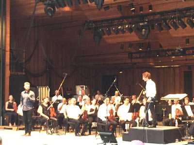 Composer Samuel Adams introduces his composition, Radial Play to the Britt Festivals audience, as Maestro Teddy Abrams and the Britt Orchestra look on, at Britt Pavilion in Jacksonville, OR on Aug. 1, 2015.